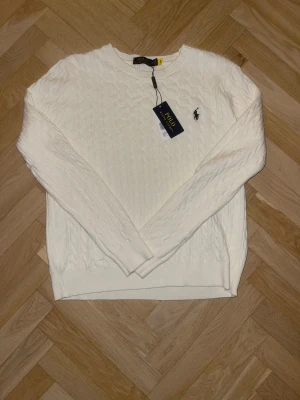 Vit stickad tröja Polo Ralph Lauren - Vit stickad tröja från Polo Ralph Lauren med klassiskt kabelstickat mönster och rund hals. Tröjan har långärmade ärmar och den ikoniska Polo-loggan broderad på bröstet. Perfekt för dig som gillar stilrena och tidlösa plagg.