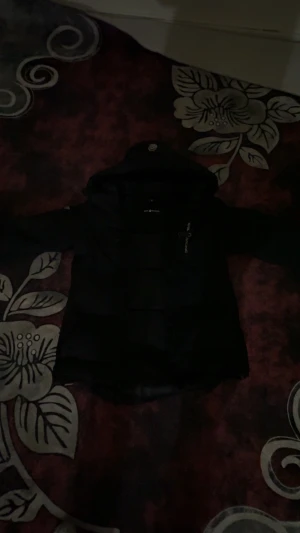 Svart Sail Racing pufferjacka med huva - Svart pufferjacka från Sail Racing med huva och coola vita logodetaljer på ärmar och bröst. Jackan har dragkedja framtill och en extra ficka med dragkedja på bröstet. Perfekt för dig som vill ha en varm och snygg jacka med streetkänsla.