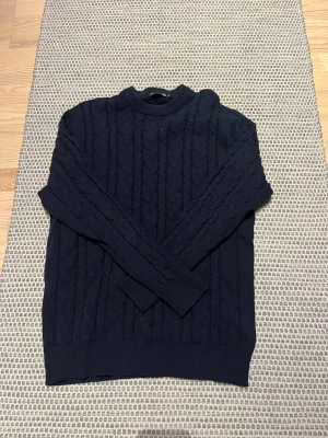 Mörkblå stickad tröja med kabelmönster - Snygg mörkblå stickad tröja med klassiskt kabelmönster och rund hals. Tröjan har långa ärmar och ribbade muddar vid ärmslut och nederkant. 