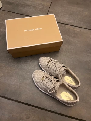 Beige sneakers från Michael Kors - Snygga beige sneakers från Michael Kors med MK-logga i guld på snörningen. Skorna har ett diskret mönster över hela ovandelen och är tillverkade i textil med vita platta sulor. Som nya, Använda 2-3 gånger endast. Storlek 38.5