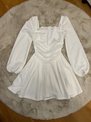 Vit kort klänning med puffärmar - helt ny - Superfin vit kort klänning med långa puffärmar och fyrkantig urringning. Klänningen har draperad midja och en lätt utsvängd kjol. Stängs med dragkedja i ryggen och är sydd i ett slätt, mjukt tyg som ger en elegant känsla.