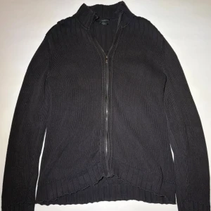 Svart stickad kofta från Calvin Klein - Svart stickad kofta från Calvin Klein Jeans med ribbad struktur och dragkedja framtill. Hög krage och långa ärmar, samt snygga axelknappar och logotyp på vänster ärm. Perfekt för dig som gillar stilrena och klassiska plagg.