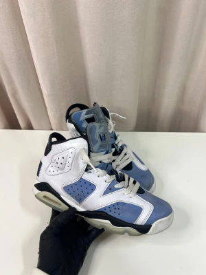 Nike Air Jordan 6 - Nike Air Jordan 6 sneakers i blått och vitt med svarta detaljer. Skorna har snörning, perforerade paneler och en platt sula med synlig luftdämpning. Materialet är en mix av skinn och syntet, och tungan har ett unikt mönster i blått och grått. Perfekt för dig som gillar klassiska basketmodeller.