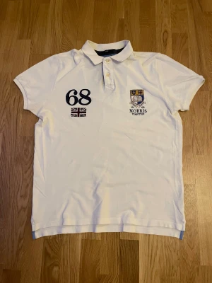 Vit pikétröja Morris Tradition L - Snygg vit pikétröja från Morris Tradition i storlek Large. Tröjan har broderade detaljer med siffran 68 och brittisk flagga på bröstet samt ett stilrent emblem med texten 'Morris Tradition'. Klassisk krage och knappar framtill. Perfekt för en preppy look.