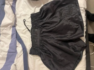 Svarta Muay Thai shorts från Venum - Säljer ett par svarta Muay Thai shorts från Venum i storlek L. De har högmidjad passform med bred resår och snörning i midjan. Materialet är glansigt syntet och har Venum-loggan i svart på framsidan. Perfekta för träning och kampsport.
