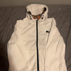 Vit Burberry vindjacka med huva - Säljer en fräsch vit vindjacka från Burberry med klassiskt rutigt foder i huvan. Jackan har svart dragkedja, snörning vid huvan och Burberrys ikoniska logga på bröstet. Perfekt för dig som vill ha en clean och stilren look. Storlek S/XS