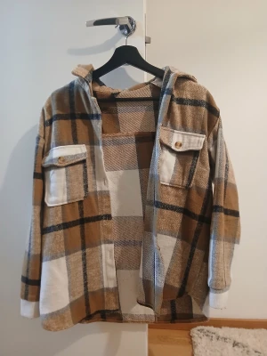 Rutig overshirt från SHEIN brun/vit - Overshirt från SHEIN i brun, vit och svart rutig design. Skjortan har huva, två stora bröstfickor med knappar och en loose passform. Perfekt lager-på-lager-plagg med mjukt material och avslappnad vibe.