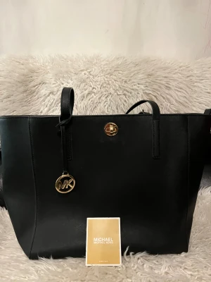 Svart axelväska från Michael Kors - Stilren svart axelväska från Michael Kors i skinn med guldiga detaljer och MK-hänge. Väskan har en fyrkantig form, långa handtag och ett snyggt vridlås framtill. Perfekt storlek för att få plats med allt du behöver och passar till många olika looks.ÄVEN HELT NY ALDRIG ANVÄND