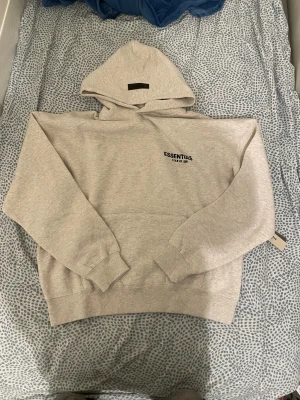 Grå Essentials Fear of God hoodie - Snygg ljusgrå hoodie från Essentials Fear of God med svart logotyptryck både fram och bak. Tröjan har huva, ribbade muddar och en klassisk känguruficka. Perfekt för dig som gillar streetwear och vill ha en clean look.