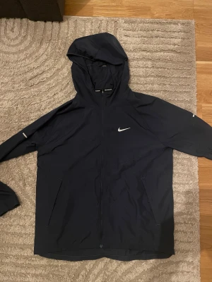 Svart Nike vindjacka med huva - Nike vindjacka i svart med stilren design och huva. Jackan har dragkedja framtill, två sidofickor och reflekterande detaljer på ärmarna. Perfekt för löpning och utomhusaktiviteter. Materialet är lätt och syntetiskt, vilket gör den smidig att bära.