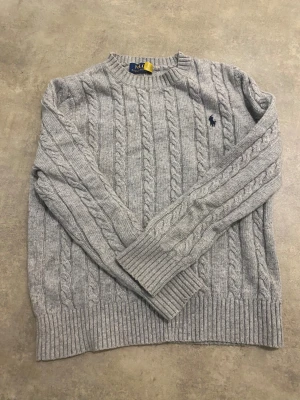 Grå stickad tröja från Polo Ralph Lauren - Säljer en grå stickad tröja från Polo Ralph Lauren med klassiskt kabelstickat mönster och rund hals. Tröjan har långa ärmar och ribbade muddar vid ärmslut och nederkant. Diskret Polo-logga broderad på bröstet. Perfekt för dig som gillar stilren och chill look. Det är storlek s men den kan va liten i storleken 