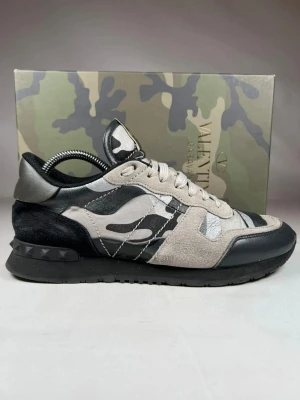 Valentino Garavani Rockrunner sneakers - Säljer ett par Valentino Garavani Rockrunner sneakers i svart, grått och beige med camouflage-mönster. Skorna har coola nitar på hälen, snörning och mix av mocka och skinn. Perfekta för dig som vill ha en unik och lyxig sneaker med street vibe.