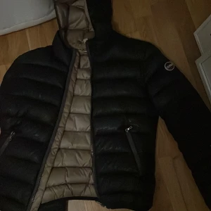 Svart jacka från colmar - Säljer en svart pufferjacka från colmar med beige insida och huva. Jackan har horisontella quiltade paneler, två fickor med dragkedja och en snygg logotyp på ärmen. Perfekt för dig som vill ha en varm och stilren jacka