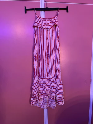 Randig långklänning med volang - Långklänning med smala axelband och volang upptill. Klänningen har röda och vita vertikala ränder och en bred volangkant nertill med horisontella ränder. Luftig och flowy modell, perfekt för varma dagar.