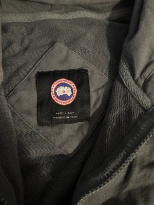 Mörkgrön  hoodiejacka från Canada Goose - Snygg mörkgrå hoodiejacka från Canada Goose med dragkedja framtill, huva och ribbade muddar. Jackan har broderad logga på ärmen och är tillverkad i mjuk bomull. Perfekt för dig som vill ha en stilren och bekväm jacka med streetkänsla.