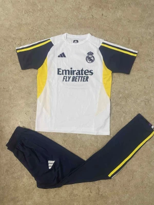 Real Madrid fotbollströja Adidas - Snygg Real Madrid fotbollströja från Adidas i vitt med mörkblå och gula detaljer. Kortärmad modell med klubbmärke och Emirates Fly Better-tryck på bröstet. Tillverkad i lätt och ventilerande material, perfekt för träning eller match.