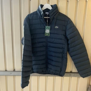 Mörkblå dunjacka från Lacoste M - Snygg mörkblå dunjacka från Lacoste i klassisk passform. Jackan har quiltad design, dragkedja framtill och det ikoniska Lacoste-krokodilmärket på bröstet. Perfekt för kyliga dagar och enkel att matcha med din stil.