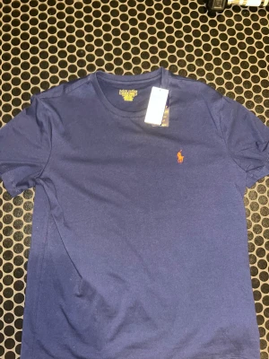 Blå t-shirt från Polo Ralph Lauren - Snygg marinblå t-shirt från Polo Ralph Lauren med klassisk rund hals och orange broderad logga på bröstet. Modellen är slim fit och tillverkad i mjuk bomull som känns skön mot huden. Perfekt för dig som gillar stilrena och ikoniska plagg. Priset kan diskuteras bara höra av er vid frågor/funderingar