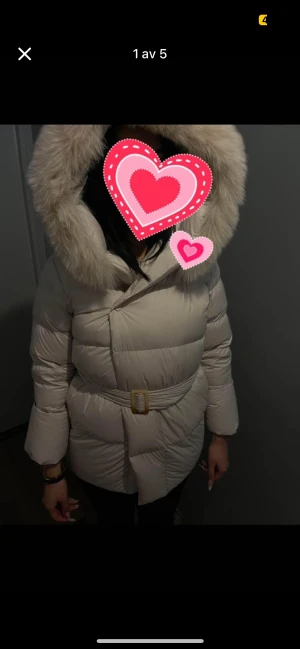 Beige dunjacka med pälsluva och bälte - Säljer en beige dunjacka med stor fluffig pälsluva och bälte i midjan. Jackan är quiltad och har en snygg, figurnära passform. Perfekt för kalla dagar med sin tjocka och varma design. Långa ärmar och knäppning framtill.