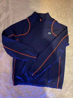 Marinblå vindjacka från Lacoste - Snygg marinblå vindjacka från Lacoste med orange detaljer längs sömmarna och klassisk krokodillogga på bröstet. Jackan har hög krage, hel dragkedja och elastiska muddar. Perfekt för dig som gillar sportig stil och vill sticka ut med coola färgkontraster.