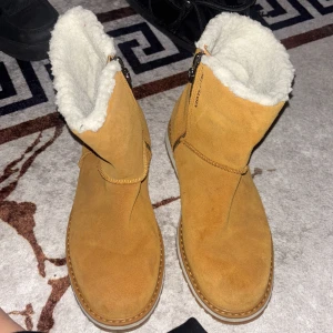 Helly Hansen beige boots med foder - Säljer ett par beige boots från Helly Hansen i storlek 38. Skorna har mjukt vitt foder på insidan, mockaliknande utsida och grov gummisula med HH-logga. Dragkedja på sidan för enkel påtagning. Perfekta för kalla dagar och har en rund tå.
