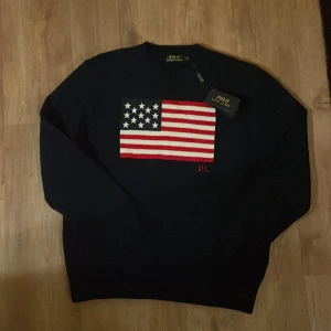 Mörkblå stickad tröja Polo Ralph Lauren - Snygg mörkblå stickad tröja från Polo Ralph Lauren med amerikansk flagga broderad på bröstet och RL-detalj i rött. Rund hals med ribbad kant och långa ärmar. Perfekt för dig som gillar klassisk och sportig stil.