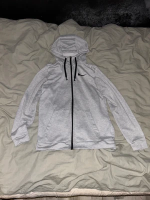 Grå hoodiejacka från Nike - Snygg grå hoodiejacka från Nike med svart dragkedja och svarta snören i huvan. Jackan har en klassisk Nike-logga på bröstet och två praktiska fickor framtill. Perfekt för chill eller sportiga dagar.