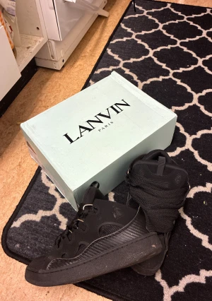 Svarta sneakers från Lanvin Paris - Lanvin Paris sneakers i helsvart med chunky design och breda snören i fiskbensmönster. Skorna har rund tå, platt sula och detaljer i både skinn och mesh. Metallöglor och diskret Lanvin-logga på sidan ger en lyxig vibe. Perfekt för dig som gillar statement-sneakers.