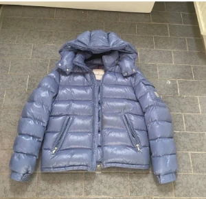 Moncler Maya Ljusblå Dunjacka  - Säljer en ljusblå Moncler-dunjacka med avtagbar huva. Jackan är lätt, varm och sitter riktigt bra. Den har dragkedja fram, två sidofickor med dragkedja samt en ficka på ärmen.  Storlek: Passar dig som är runt 175 cm och 70 kg (för referens är det jag på bilden).  Jackan har några små fläckar, men de är knappt synliga om man inte tittar nära .  Självklart hundra procent äkta.