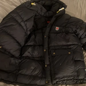 Svart pufferjacka från Fjällräven - Säljer en svart pufferjacka från Fjällräven i storlek S. Jackan har en klassisk puffad design, stora fickor med knappar och dragkedja, samt Fjällrävens ikoniska logga på bröstet fram. Priset kan diskuteras!!! Den är den fluffigaste modellen.