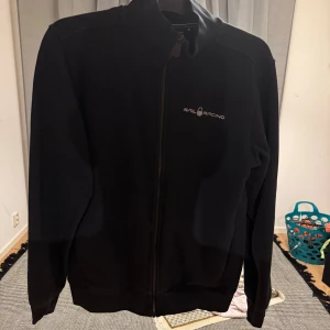 Svart Sail Racing ziptröja - Svart ziptröja från Sail Racing med diskret logga på bröstet och en cool detalj i nacken. Tröjan har hög krage, hel dragkedja och långa ärmar. Perfekt för dig som gillar stilren design och sportiga vibbar. Den kostar för 1300kr ifrån affären men säljer den nu för 250kr bara för att den är lite sliten.