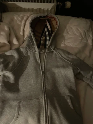 Grå hoodie från Burberry med dragkedja - Säljer en grå hoodie från Burberry med klassiskt rutigt foder i huvan. Tröjan har dragkedja framtill, två fickor och långa ärmar. Perfekt för dig som vill ha en stilren och lyxig look med ikoniska Burberry-detaljer.