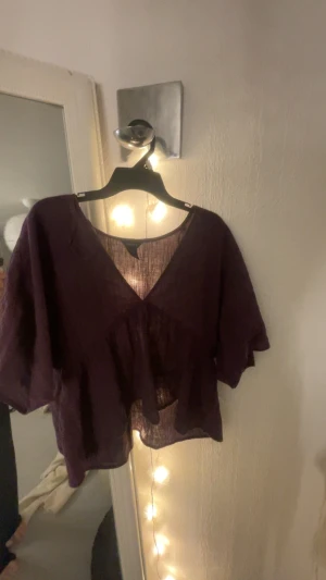 Lila blus med v-ringning från LINDEX - Säljer en lila blus från LINDEX med v-ringad hals och korta, vida ärmar. Blusen har en luftig och lätt struktur i ett transparent tyg, samt en volangdetalj framtill. Perfekt för dig som gillar avslappnad och trendig stil.