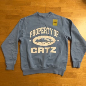 Blå sweatshirt från Corteiz CRTZ - Blå sweatshirt från Corteiz CRTZ i storlek Large. Tröjan har ett stort vitt tryck med texten 'PROPERTY OF CRTZ' och en grafisk illustration på bröstet. Rund hals, ribbade muddar och mjukt material som känns skönt mot huden. Perfekt för dig som gillar streetwear.      Äkta, Kvitto finns!