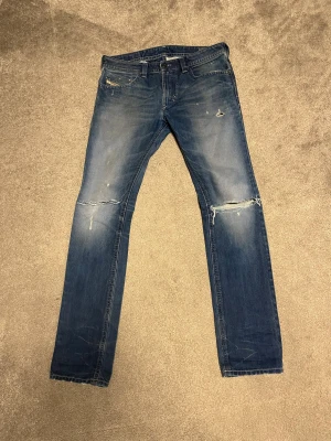 Blå slitna jeans från Diesel - Snygga blå jeans från Diesel med slitningar och hål på knäna för en cool, avslappnad look. Klassisk femficksmodell med raka ben och normal passform. Jeansen har en tvättad finish och detaljerade sömmar på baksidan. Perfekta för dig som gillar streetstyle.
