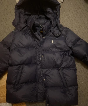Mörkblå pufferjacka från Polo Ralph Lauren - Snygg mörkblå pufferjacka från Polo Ralph Lauren med glansig finish och klassisk logga på bröstet. Jackan har huva, dragkedja och är vadderad för extra värme. Perfekt för kalla dagar när du vill hålla stilen.