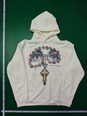 Vit hoodie Ice Valley med grafiskt tryck - Vit hoodie från Ice Valley i storlek M med ett unikt grafiskt tryck på framsidan som visar ögon, färgglada stenar och ett kors med krona. Tröjan har huva och magficka, tillverkad i mjukt syntetmaterial. Perfekt för dig som gillar statement-plagg och coola detaljer.