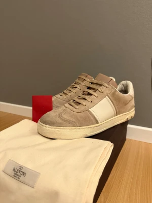 Valentino Flycrew beige sneakers - Snygga sneakers från Valentino Garavani i beige mocka med vita skinnpaneler och platt sula. Låg modell med rund tå och snörning. Märkeslogga på plösen och coola nitar på sidan av sulan. Perfekt för dig som gillar stilrena och lyxiga sneakers.