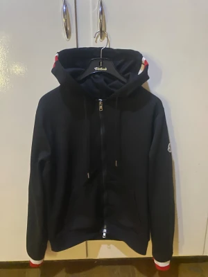 Svart hoodie från Moncler med dragkedja - Svart hoodie från Moncler med hel dragkedja, huva och snörning. Tröjan har långa ärmar med vita och röda detaljer vid mudden samt Moncler-logga på vänster ärm. Perfekt för dig som gillar stilrena och exklusiva plagg.