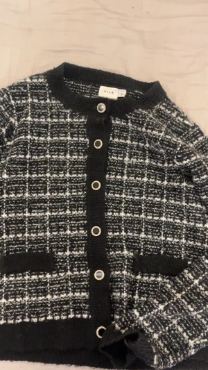 Svartvit rutig kofta från Vila - Säljer en svartvit rutig kofta från Vila i storlek M. Koftan har grovstickat material, rund halsringning och snygga dekorativa knappar framtill. Två fickor framtill och ribbade muddar vid ärmslut och nederkant. Perfekt för dig som gillar klassisk stil med en twist.
