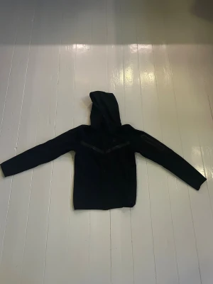 Svart Nike tech  hoodie med dragkedja - Svart hoodie från Nike med hel dragkedja framtill och huva. Snygga grå detaljer i form av två ränder över bröstet och Nike-logga på vänster sida. Tröjan har lång ärm och är gjord i mjuk bomullsmix som är skön att ha på sig. Perfekt för chill eller träning.