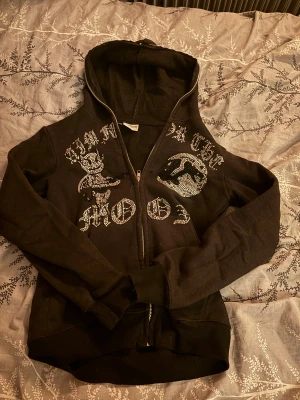 Svart hoodie Aim For The Moon XS - Svart hoodie med dragkedja från Aim For The Moon i storlek XS. Framsidan har coola paljettmotiv med text och en tecknad figur. Tröjan har huva och långa ärmar. Tröjan har tappat några stenar tyvärr men de är inget man tänker på. Pris kan diskuteras. 