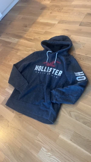 Mörkblå Hollister hoodie med tryck - Snygg mörkblå hoodie från Hollister i storlek M. Tröjan har vit snörning i huvan, stor logotyp på bröstet och tryck längs ena ärmen. Perfekt för chill dagar. 
