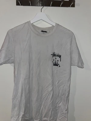 Vit Stüssy t-shirt med krontryck - Vit t-shirt från Stüssy i bomull med svart krona och logga tryckt både fram och bak. Klassisk rund hals och korta ärmar. Snygg streetwear-vibe med stor grafisk print på ryggen. Perfekt för dig som gillar bold prints och ikoniska märken. Lite småhål längst ner i ryggen men inget som tänks på vid användning 