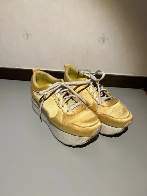 Gula plattformssneakers från W.Cobler - Unika sneakers i ljusgul/guldig satin från W.Cobler med vit plattformssula och vita snören. Skorna har rund tå och snygga detaljer i satin som ger en cool vibe. Perfekt för dig som vill sticka ut med färg och höjd på fötterna. Inga tecken på användning förutom lite smutsiga sulor!