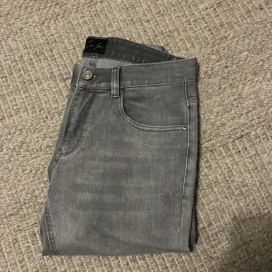 Grå slim jeans från Sacre jeans, stl 29 - Slim fit jeans från Sacre jeans i snygg grå tvätt med klassisk femficksdesign och diskret S-broderi på myntfickan. Modellen är rak och smal över benen, med knapp och dragkedja i midjan. Perfekt för dig som gillar stilrena och moderna jeans.perfekt för dig son leter efter en grisch stil. Helt nya. Nypris ligger på 1000