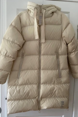 Beige lång dun jacka  - Lång beige dunjacka från Pernilla Wahlgren  med huva och breda kanaler. Jackan har dragkedja framtill, två snedställda fickor med dragkedja och ribbade snören i huvan. Perfekt för kalla dagar och ger en riktigt chill vibe.
