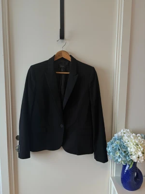 Svart kavaj från Mango Suit, stl 40 - Klassisk svart kavaj från Mango Suit i storlek 40. Snygg passform med enkla knappar framtill, spetsiga slag och fickor. Perfekt för en stilren look. Materialet är mjukt och följsamt, troligen polyesterblandning.