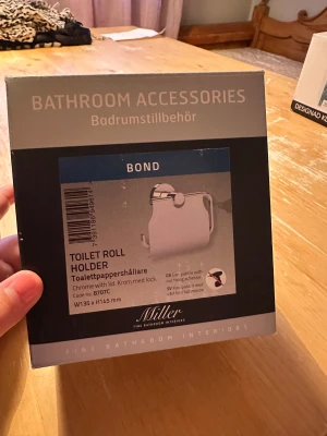 Bond Toilet Roll Holder 8707C - Elegant toalettrullehållare i krom med lock från Miller, modell Bond. Ny i förpackning, mått 135x145 mm. Perfekt för badrummet, enkel montering utan borrning möjlig. Snygg och praktisk design för en stilren badrumsinredning.