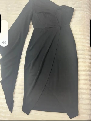 Svart oneshoulder omlottklänning - Snygg svart klänning med omlott-design och asymmetrisk oneshoulder. Klänningen har en lång ärm och en axelbandslös sida, vilket ger en cool och modern look. Passformen är figurnära och längden är kort, perfekt för dig som gillar att sticka ut.